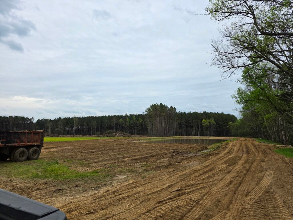 Cleared dirt field with tire tracks, a parked dump trailer at left, trees in the background, and cloudy sky