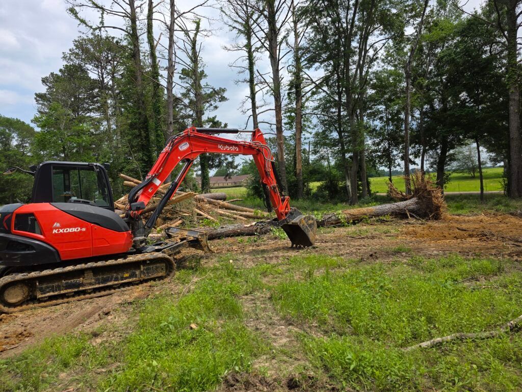 Red excavator clearing logs in a wooded area with grass and trees.
