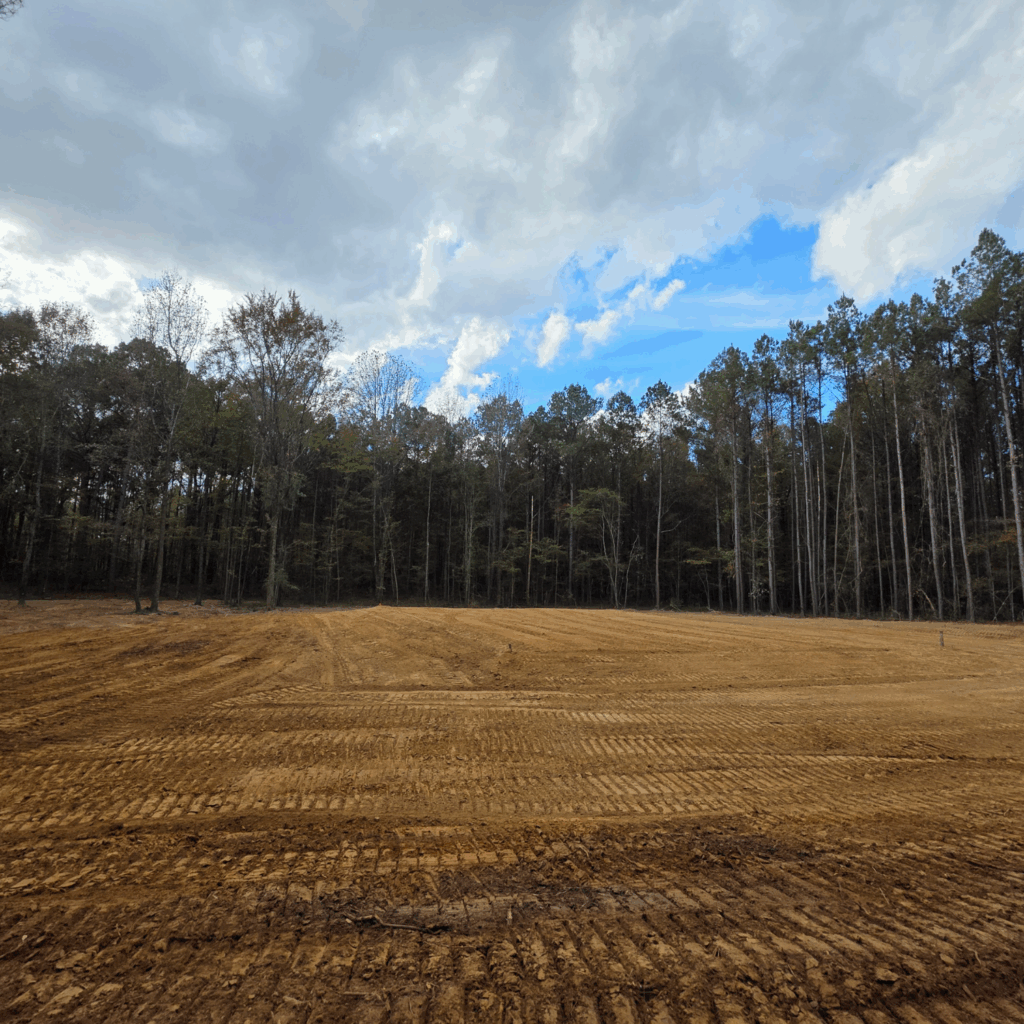 Freshly graded dirt field bordered by trees under a cloudy blue sky