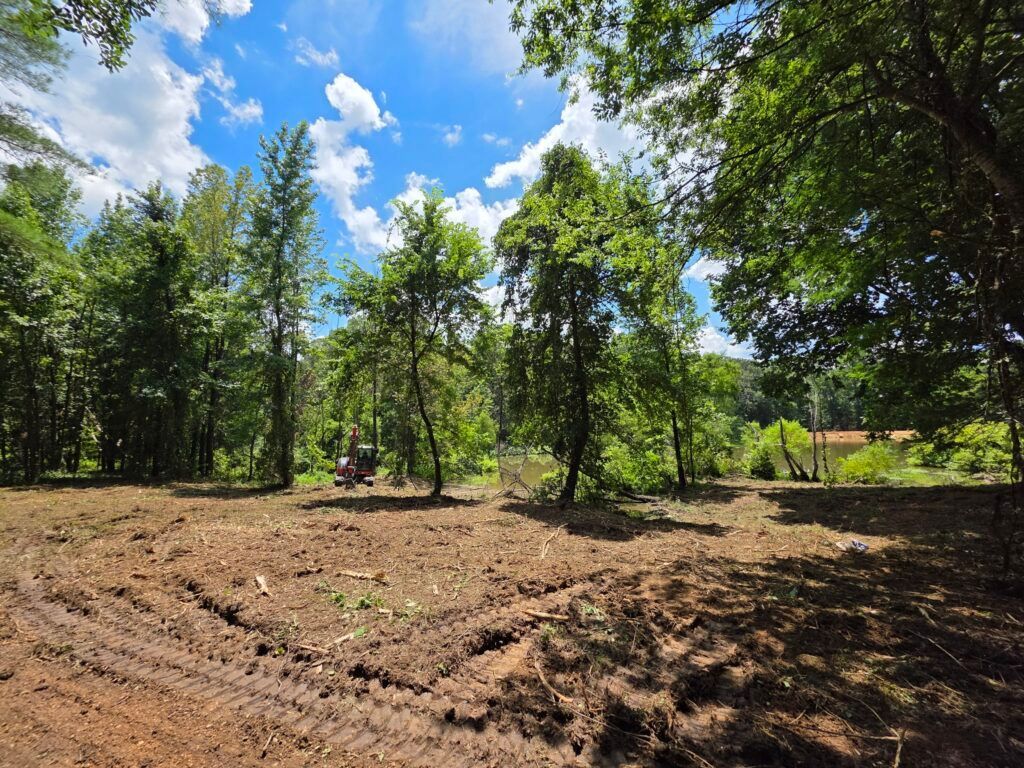 Cleared dirt lot beside trees under a bright blue sky with a small tractor in the distance