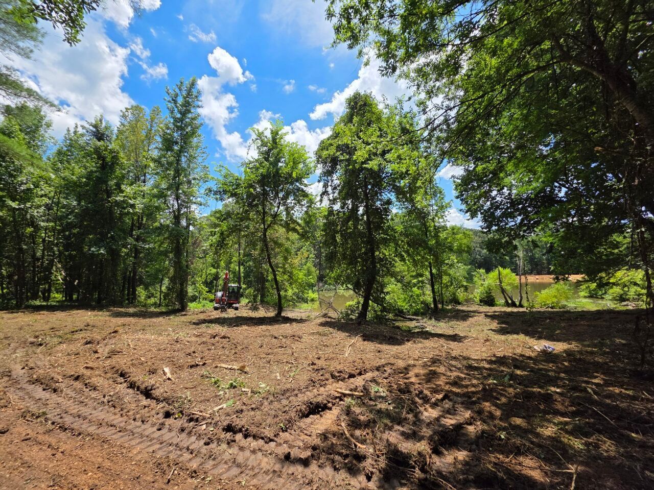 Cleared dirt path through trees under a bright blue sky with clouds, a person on a small vehicle in the distance