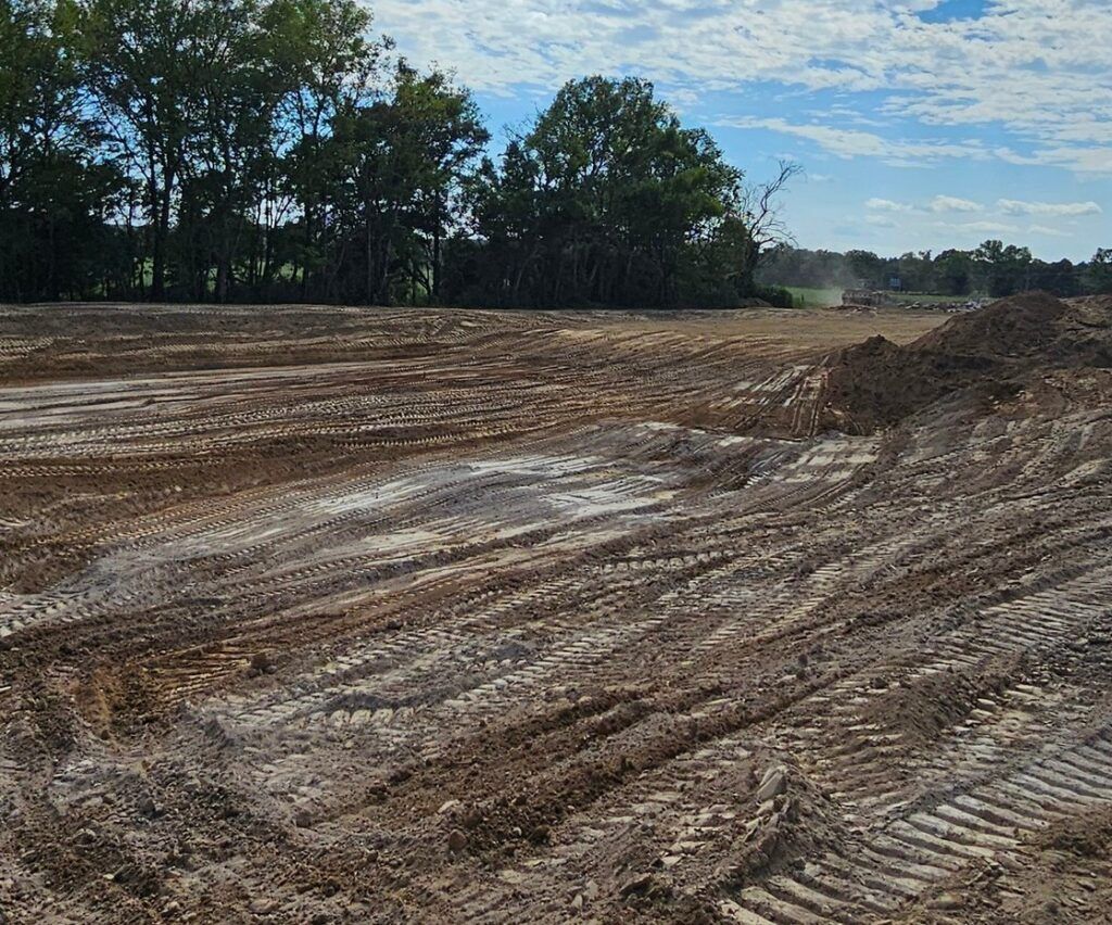Cleared dirt construction site with tire tracks, tree line, and cloudy blue sky