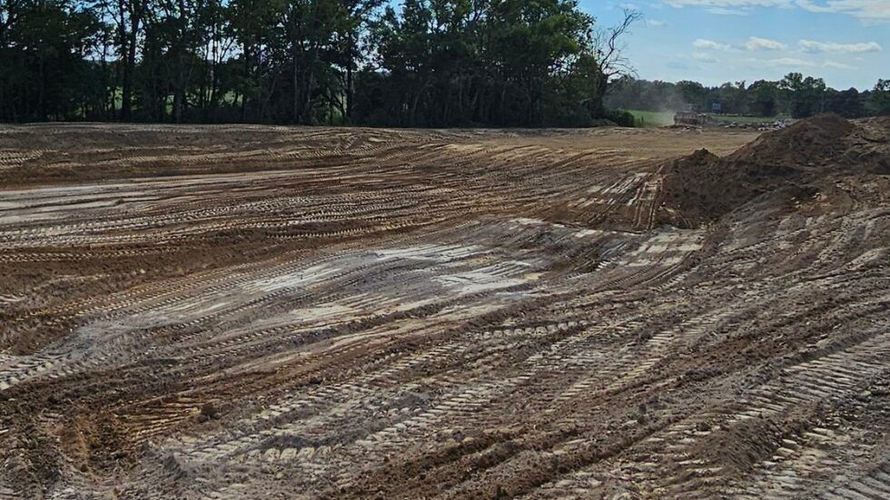 Cleared dirt field with muddy tire tracks and a pile of earth beside trees under a blue sky