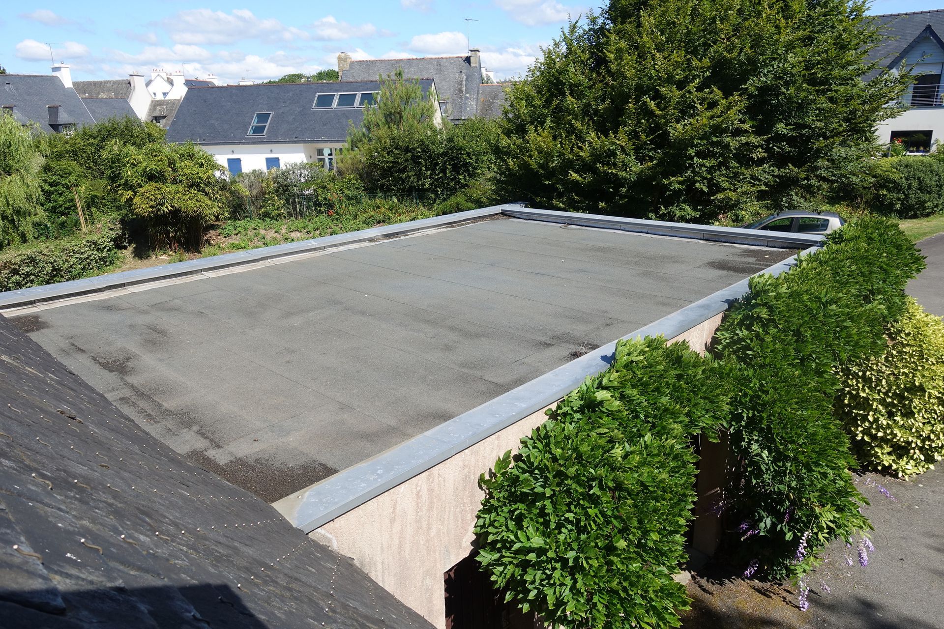 Flat roof, gray asphalt, surrounded by shrubbery and buildings in a residential area on a sunny day.