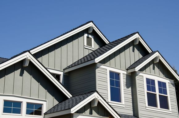 Gray house with multiple rooflines, white trim, and blue windows against a clear sky.