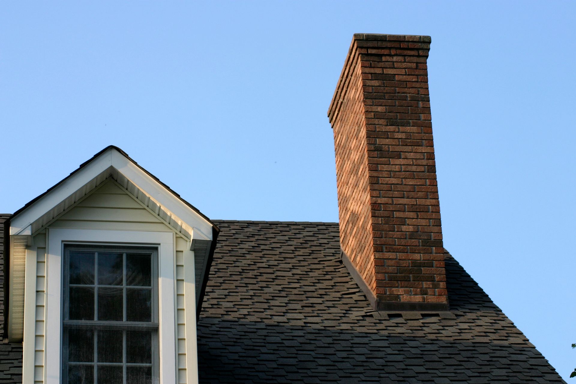 Brick chimney on a dark shingled roof next to a white-framed window against a blue sky.
