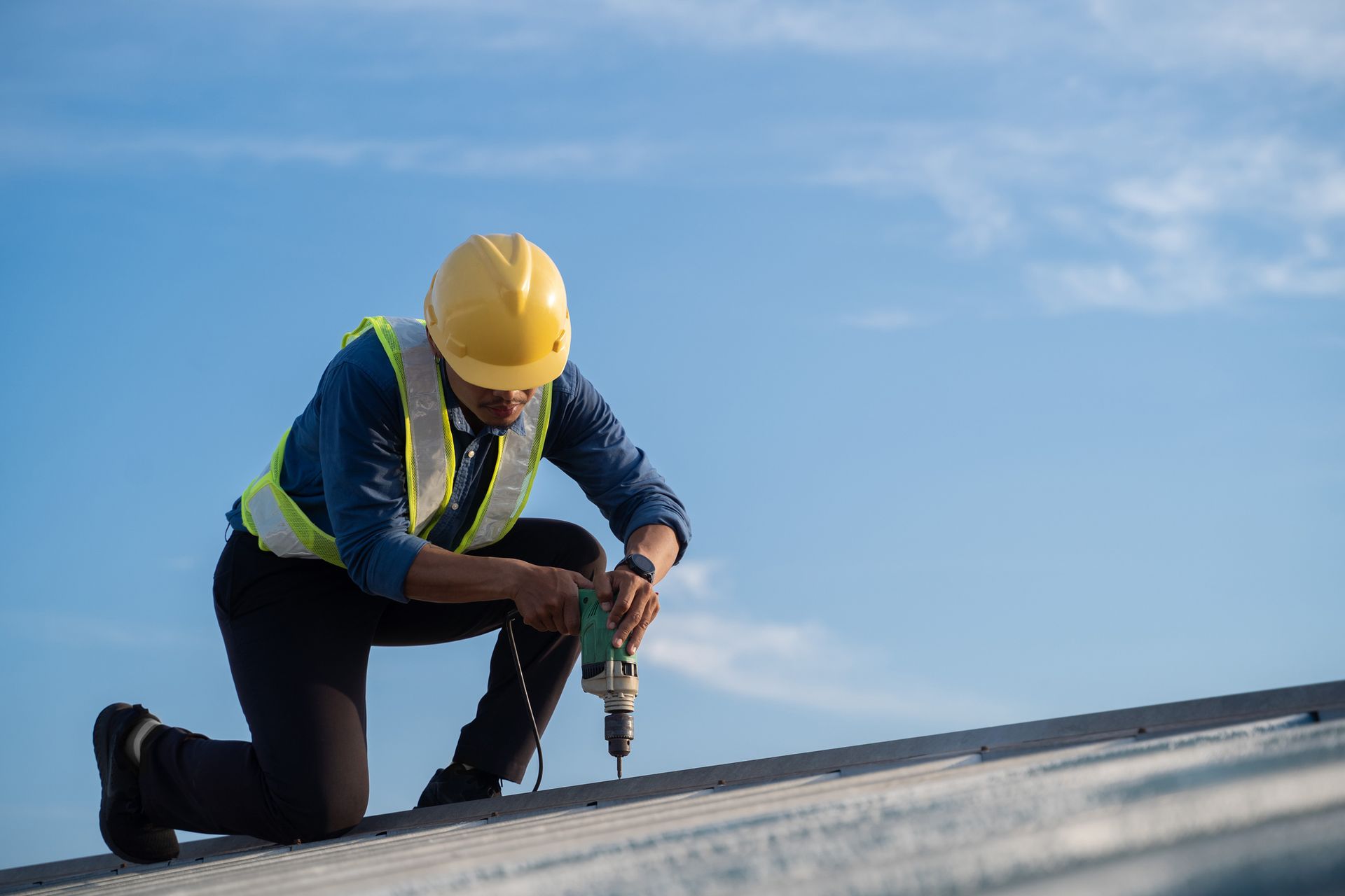Construction worker kneeling on a rooftop, using a drill. Wearing a yellow hard hat and safety vest against a blue sky.