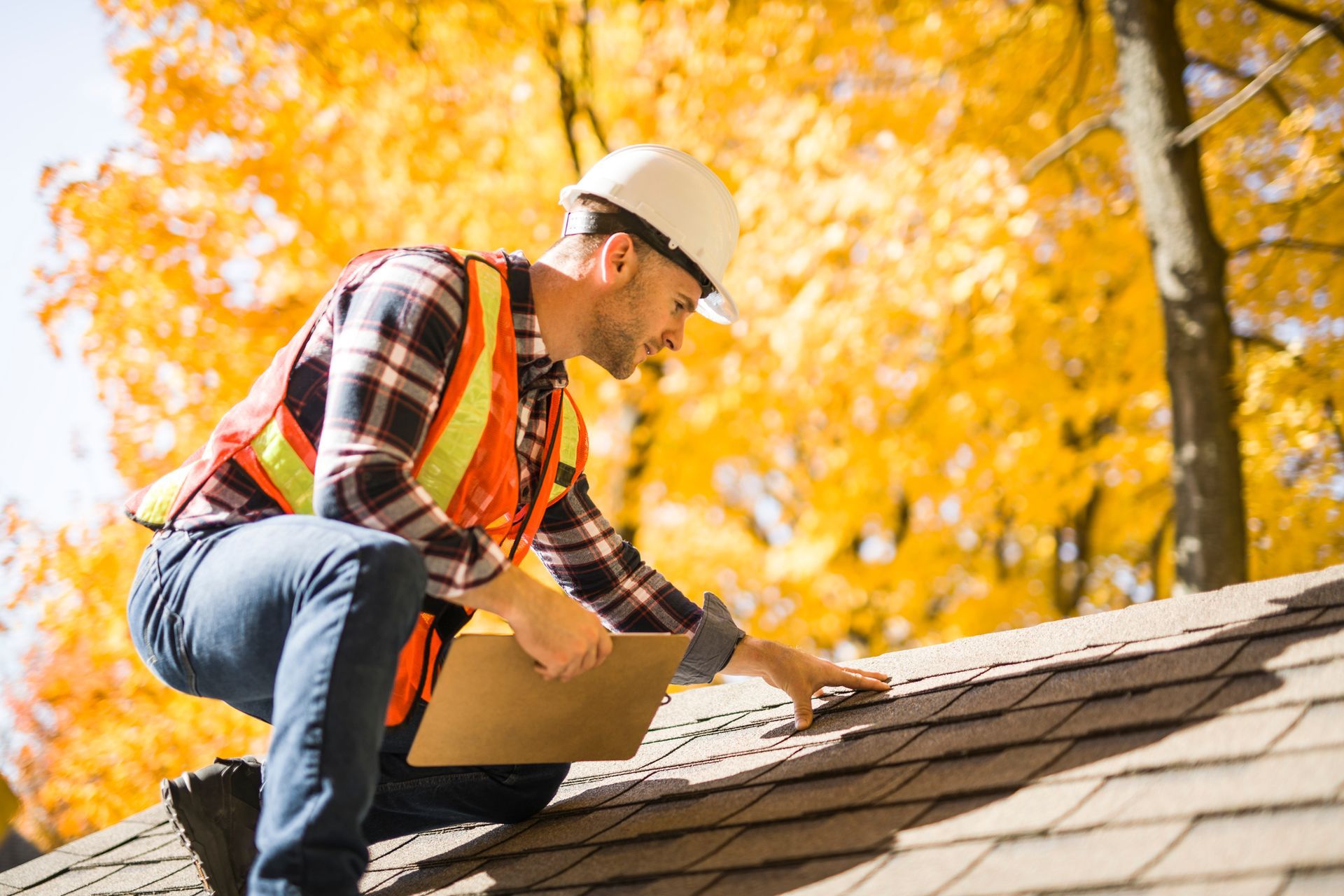 Roofer inspecting shingles on a house roof, wearing a hard hat and safety vest. Autumn foliage in the background.