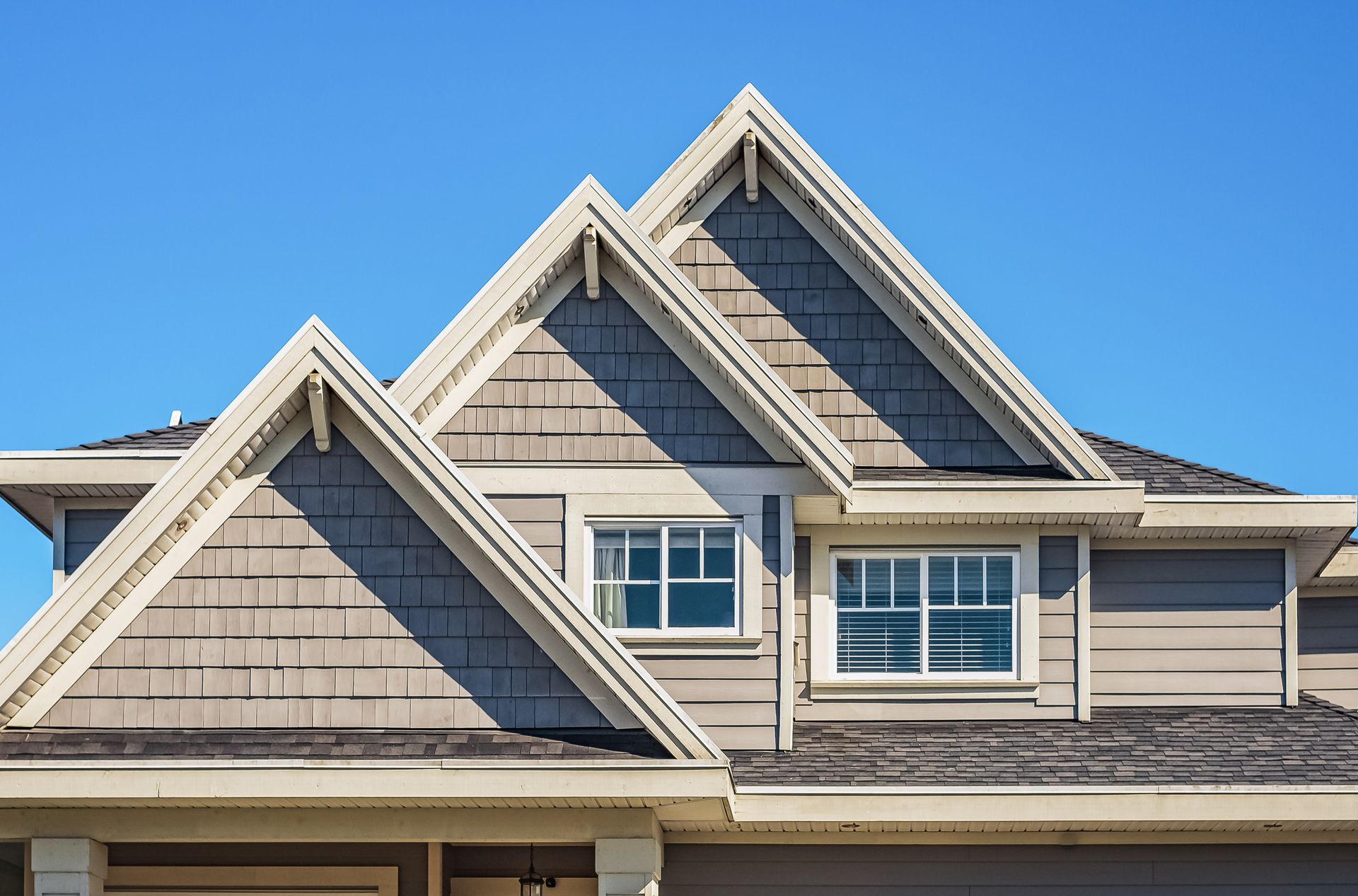 Close-up of a house roof with three gables, gray shingles, and white trim against a blue sky.
