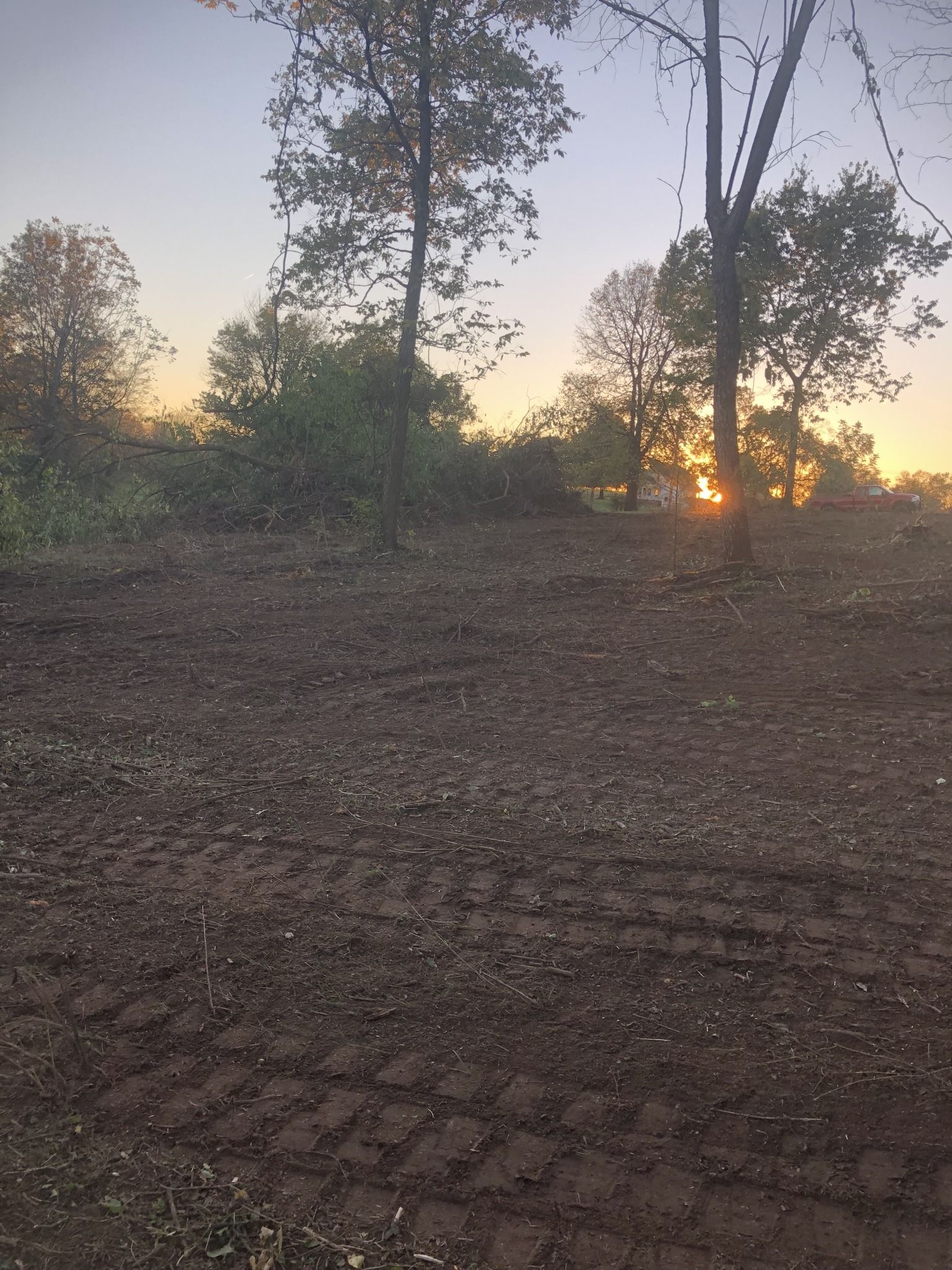 A cleared dirt field with tire tracks, trees against an orange sunset.