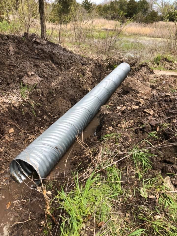 Corrugated metal pipe in a dirt trench, likely for drainage. Outdoors in a grassy field.