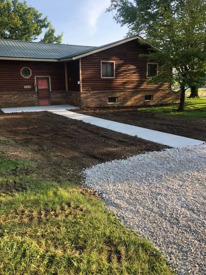 A concrete walkway leads to a cabin with a gravel driveway. Green grass surrounds it.