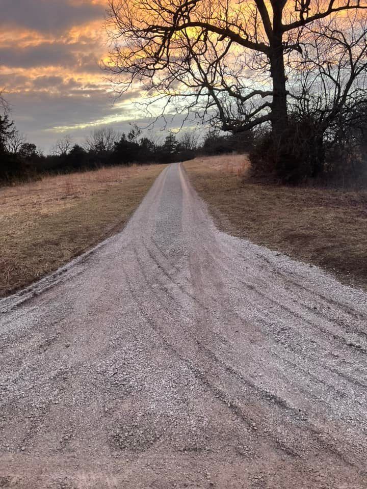 Gravel road leading toward sunset, lined with dry grass and bare trees.