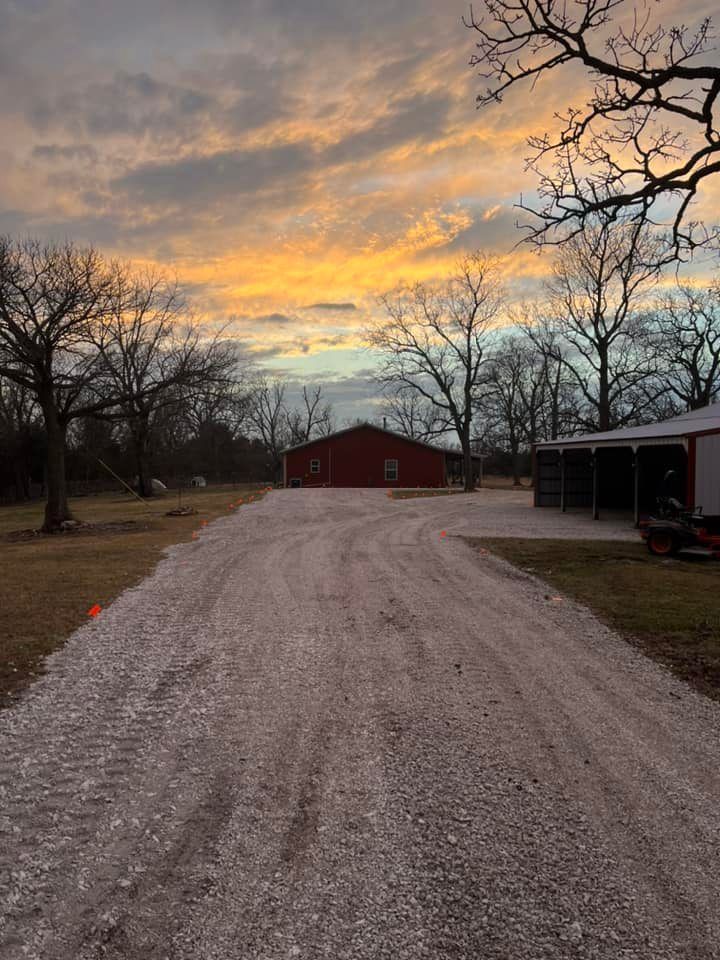 A gravel driveway leads to a red barn at sunset with an orange and yellow sky.