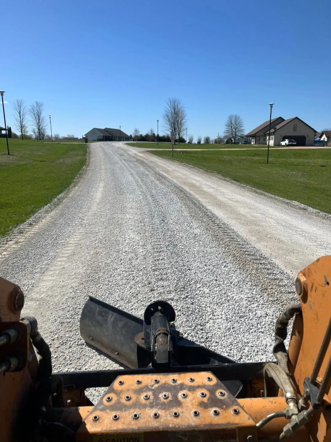 View from a tractor on a gravel driveway leading to a farmhouse under a clear blue sky