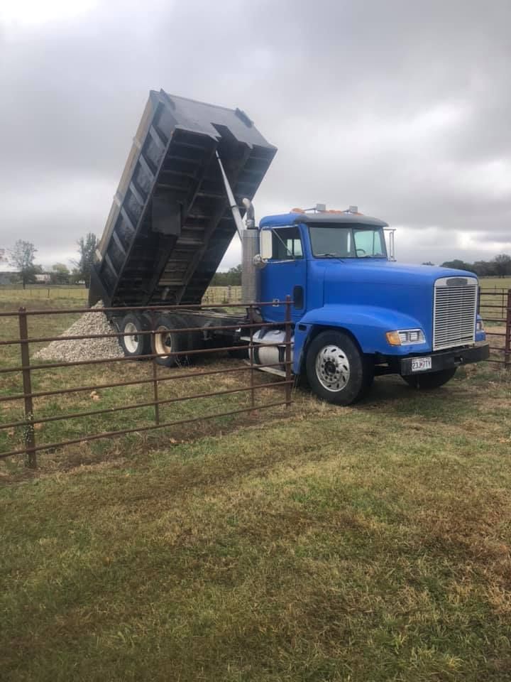 Blue dump truck unloading gravel near a brown fence on a grassy field under cloudy skies.