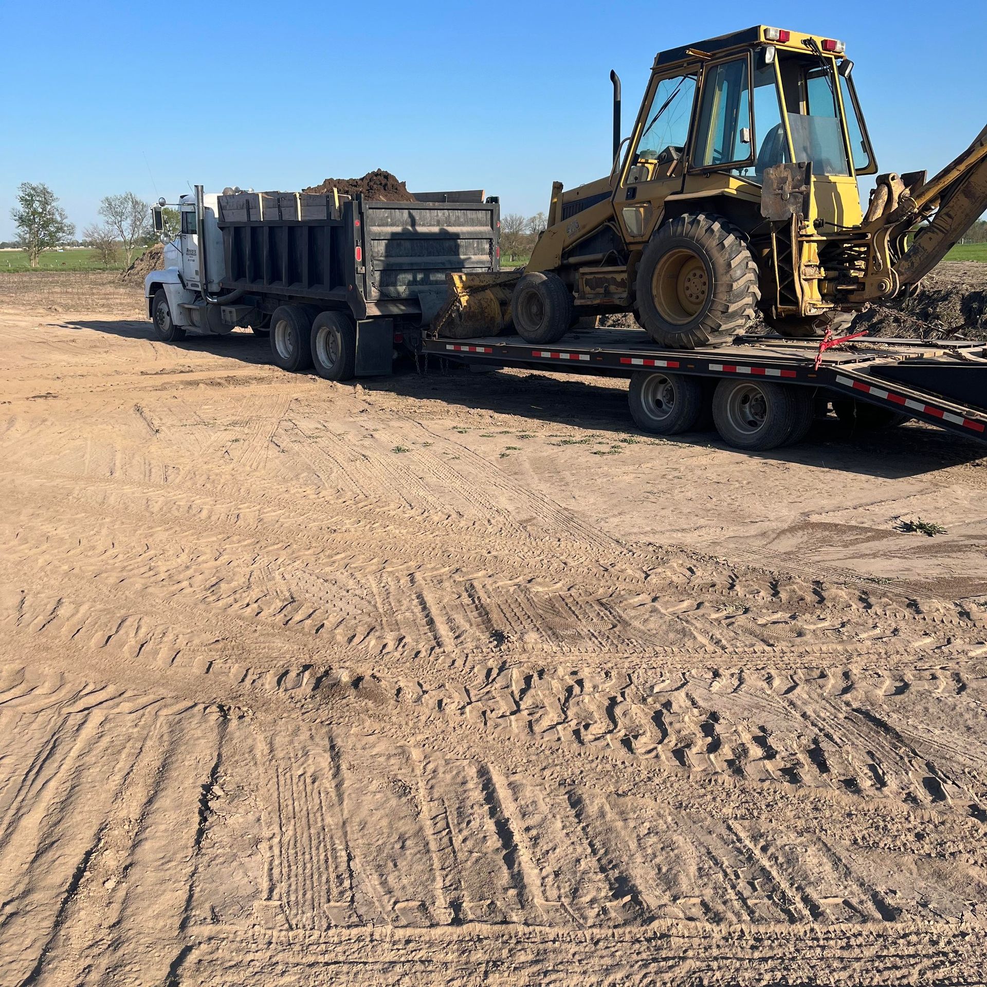 A truck and trailer transporting a yellow backhoe and a load of dirt on a brown field.