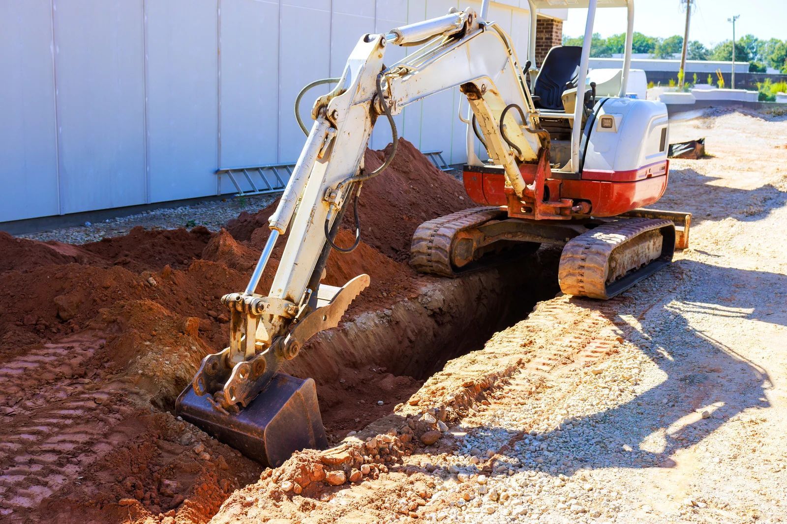 White mini excavator digging a trench beside a building on a dirt construction site