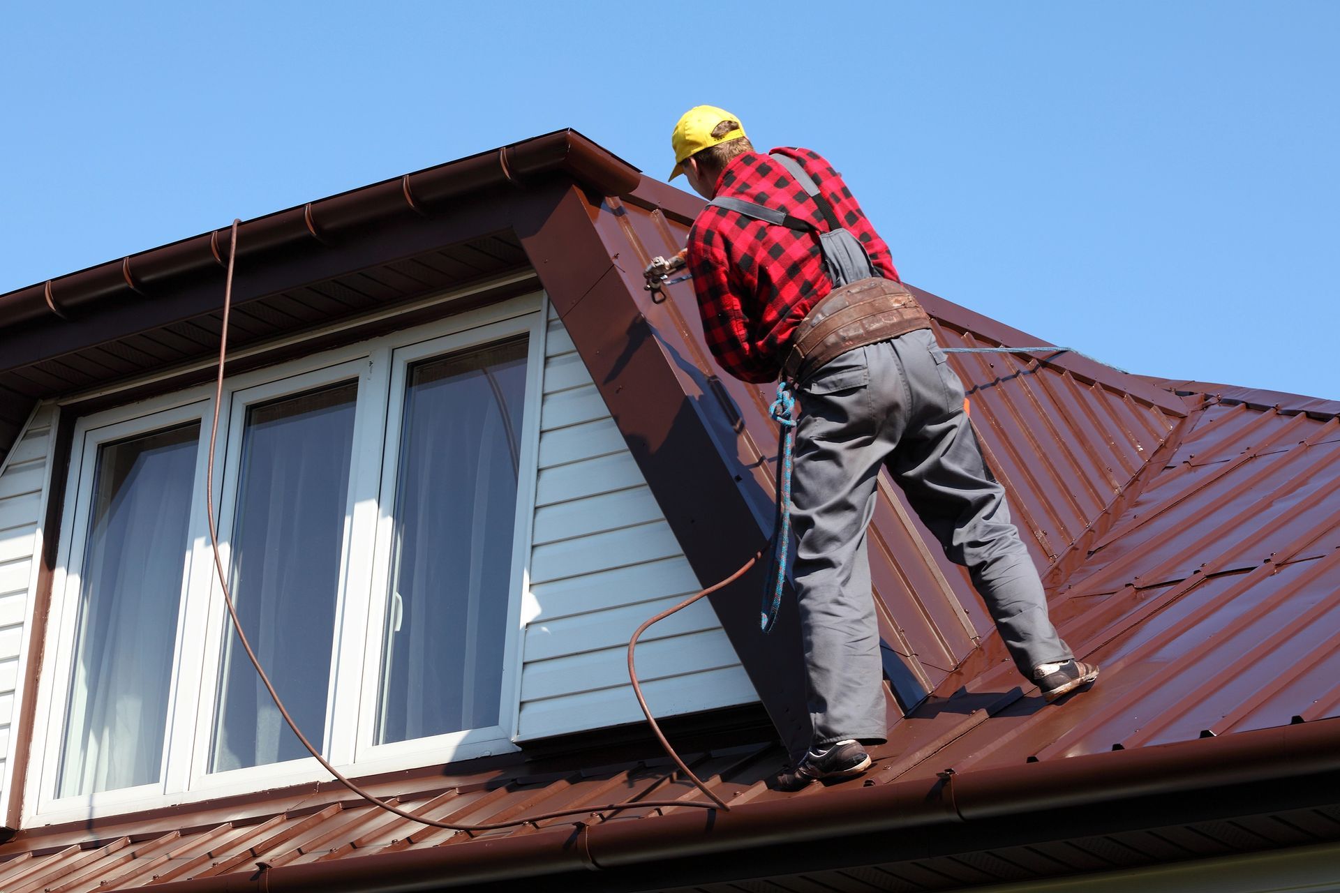 Person on a brown metal roof wearing a harness, working near a window; clear blue sky.