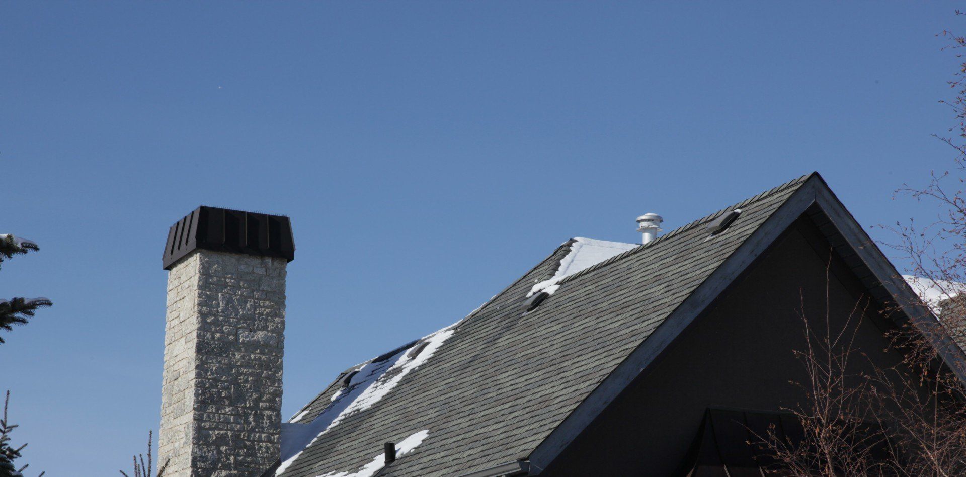Snow-dusted roof with dark shingles and a brick chimney against a clear blue sky