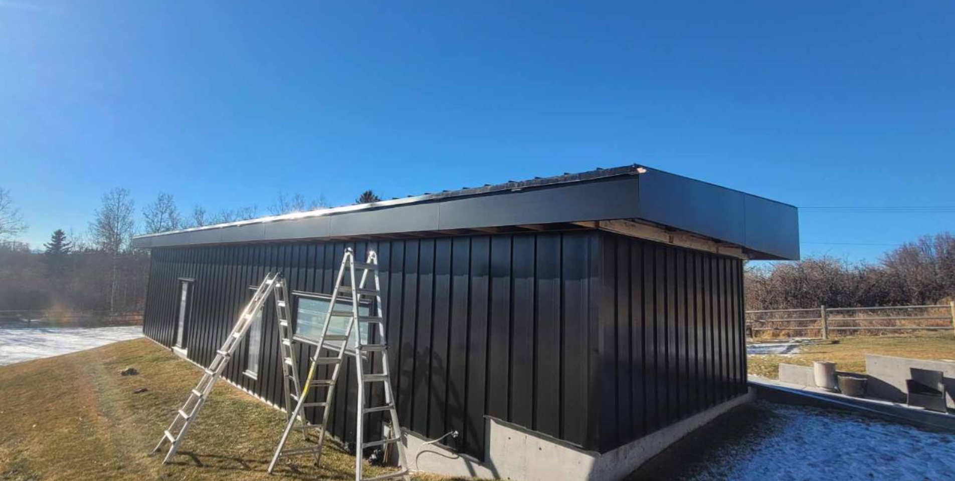 A black metal-sided building under construction with two aluminum ladders propped against the exterior.