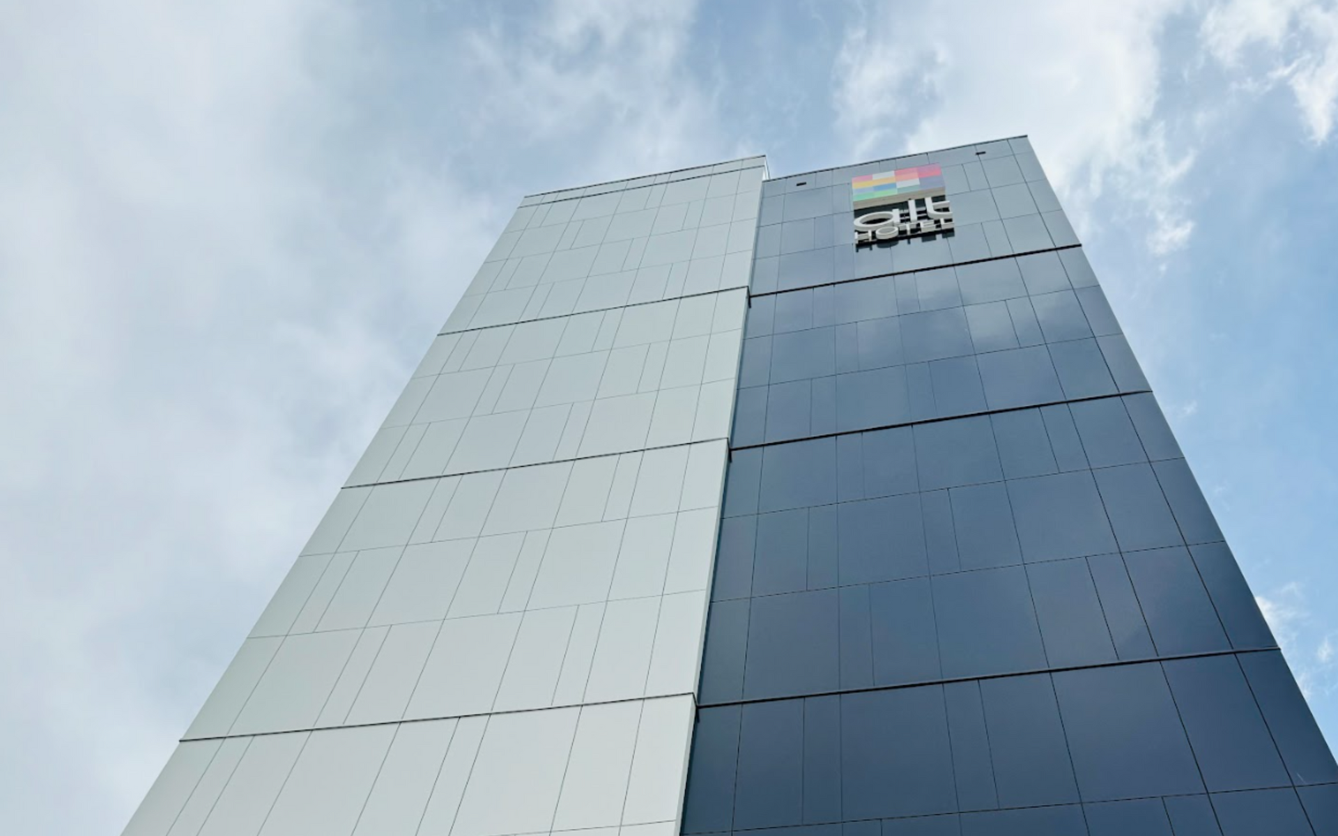 Low-angle view of a modern glass office tower against a cloudy sky, with a logo near the top.