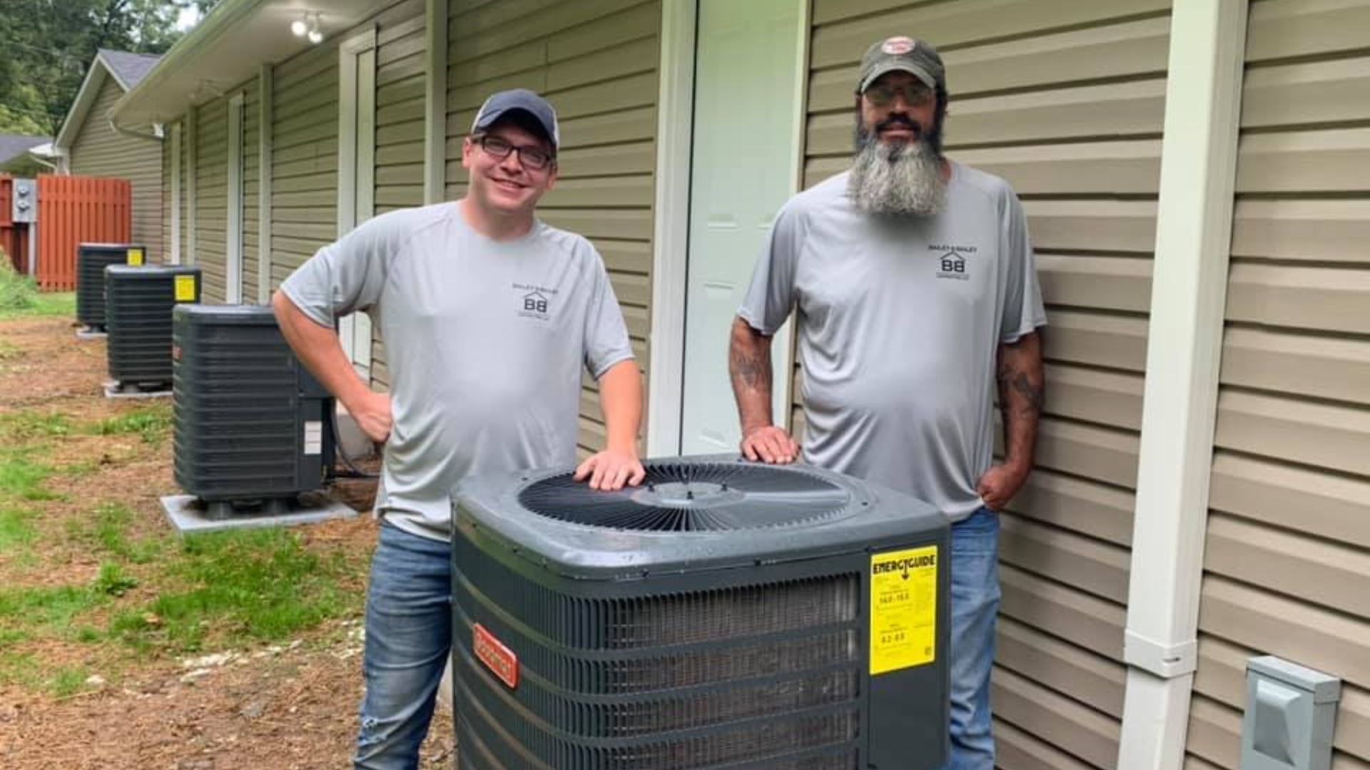 Two men in matching shirts stand by a new air conditioning unit outside a building.