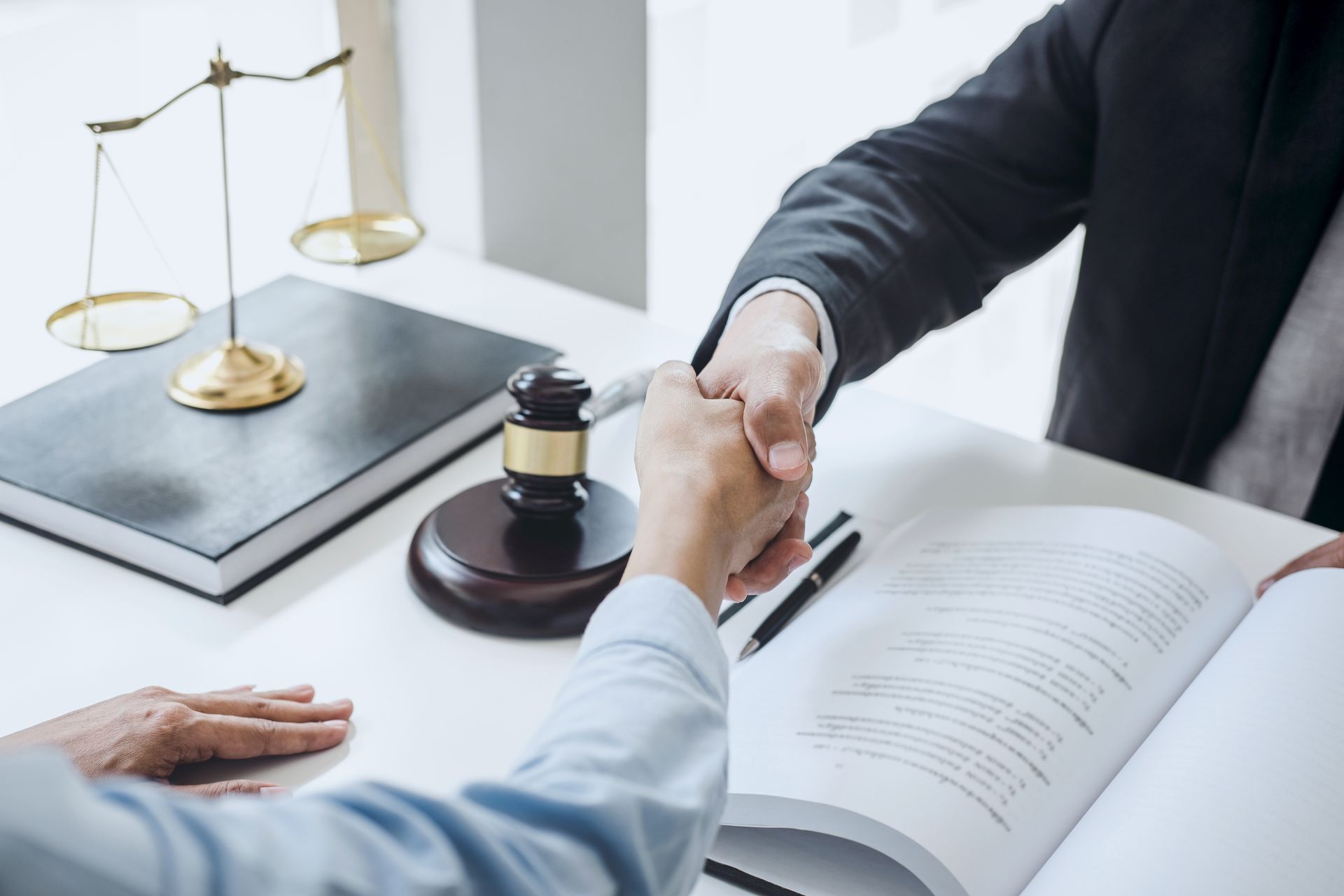 People shaking hands over an open law book, scales of justice, and a gavel on a desk.