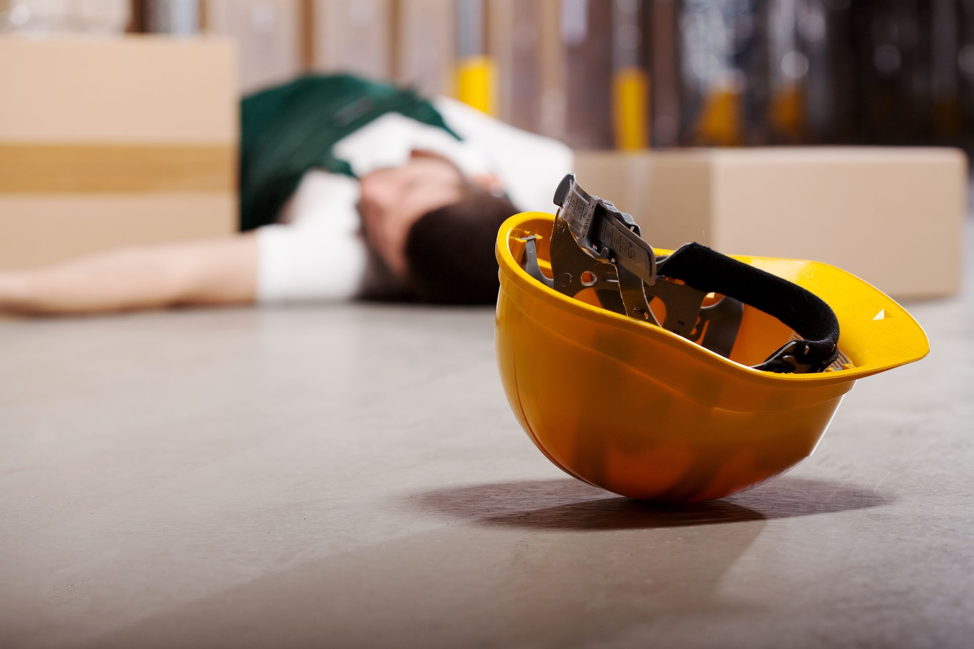 Worker lying on the floor in a warehouse, a yellow hard hat nearby.