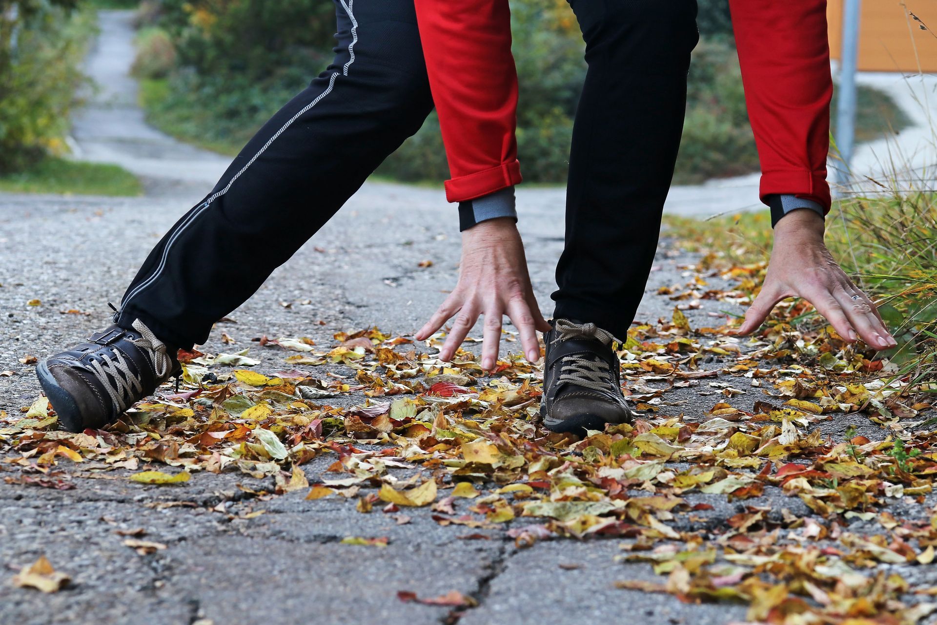 Person stretching on a leaf-covered path, hands on the ground, wearing black and red activewear.