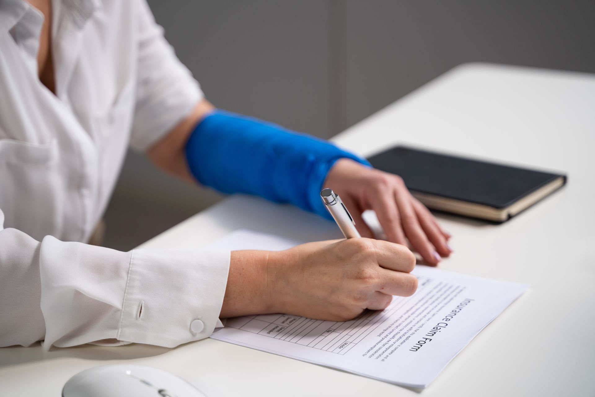 Person with blue cast on wrist signing a document at a desk with a closed notebook.