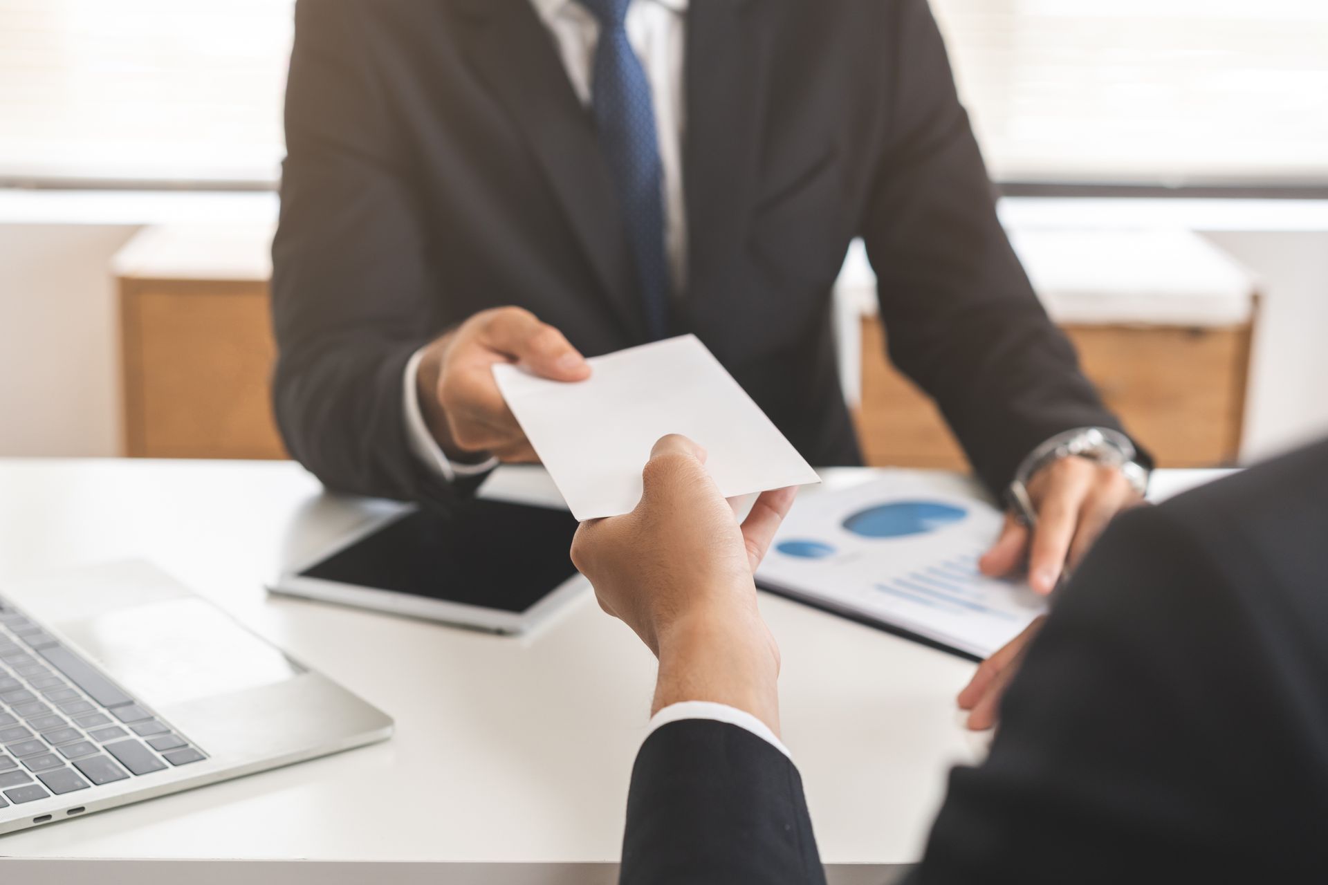 Two business people in suits exchanging a document at a desk with financial data.