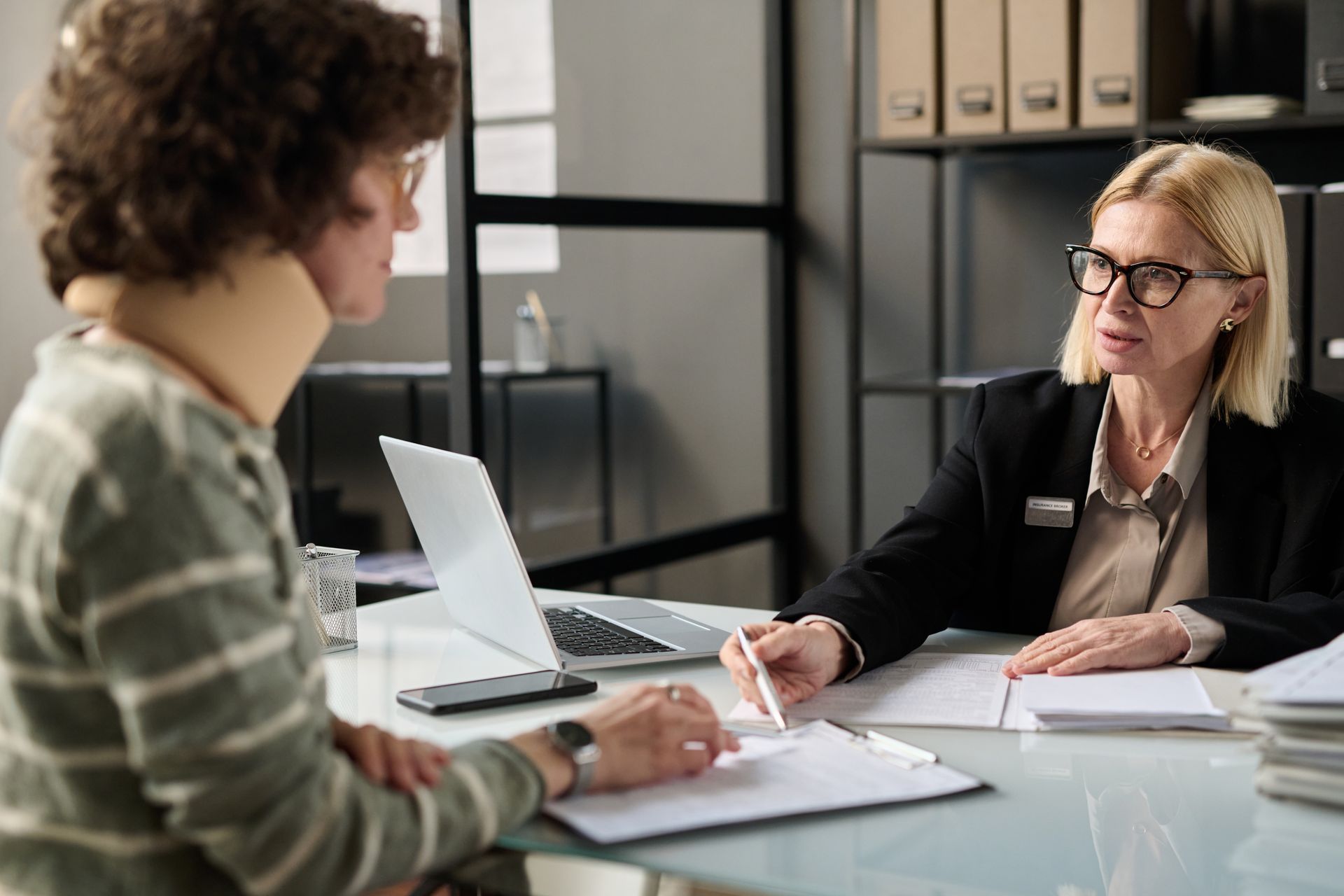 Woman in neck brace consulting with another woman at a desk with a laptop and paperwork.
