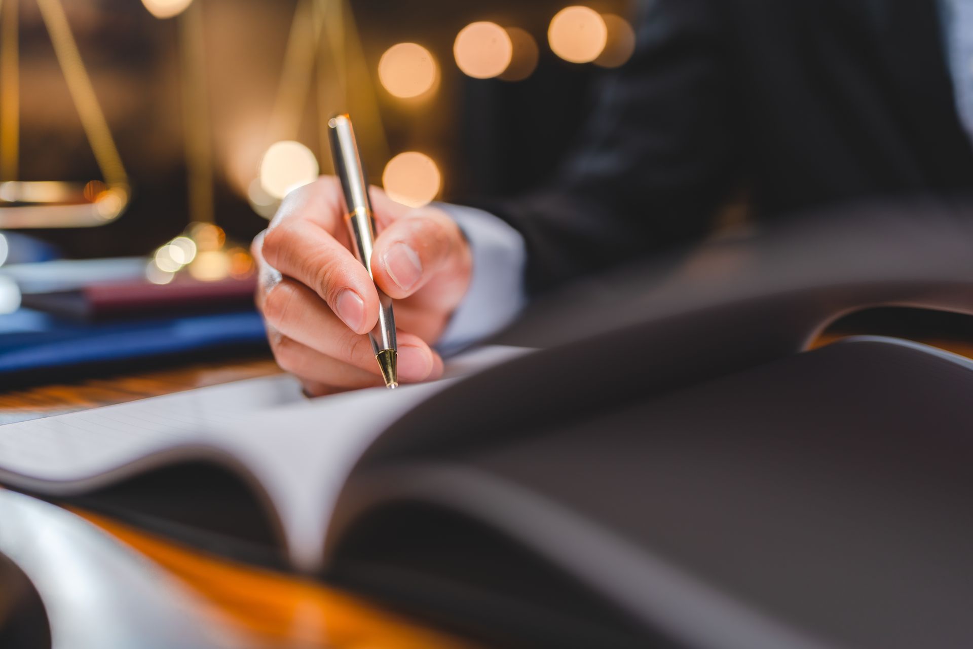 Person in suit writing in a book with a pen near a balance scale and lights.