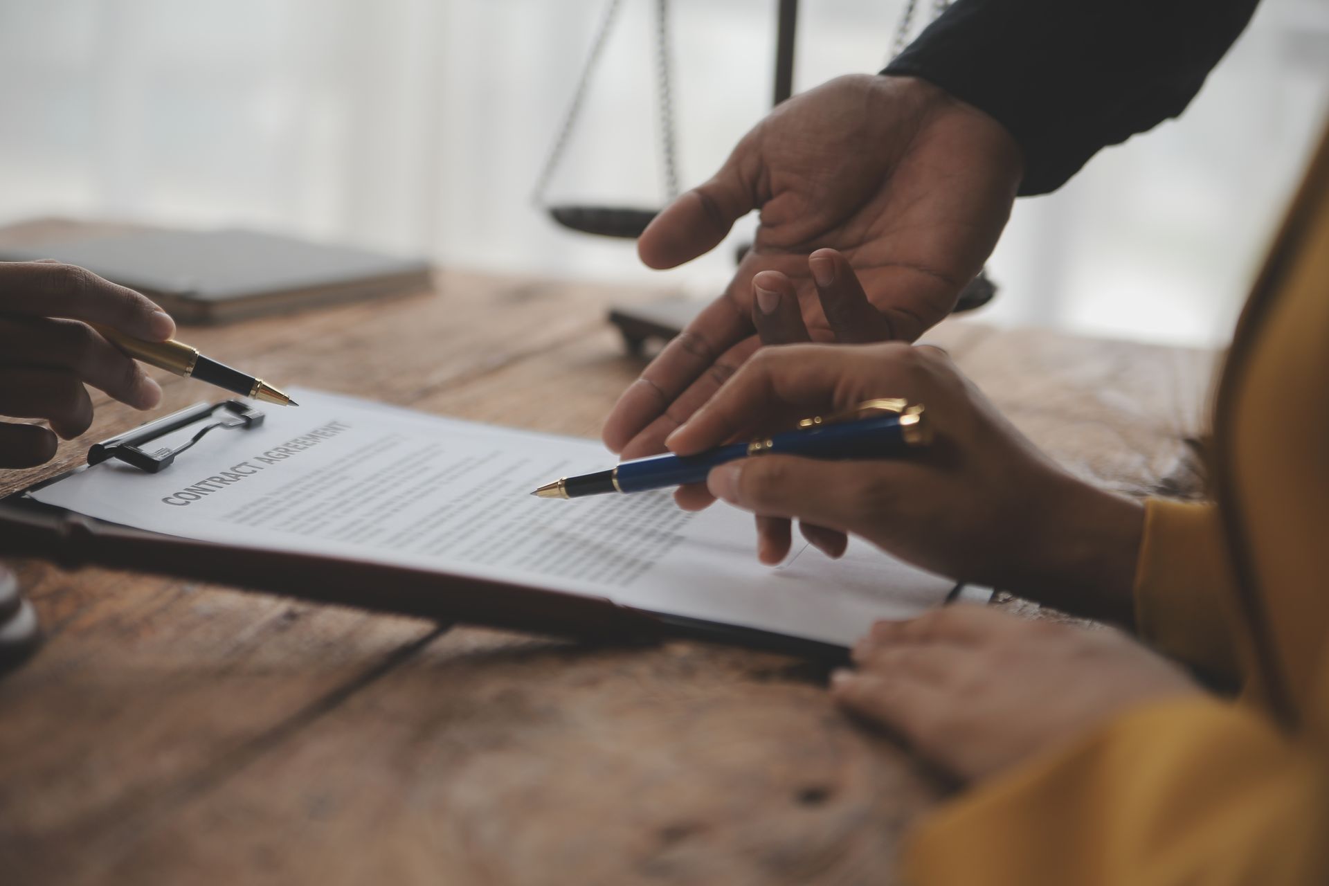 Hands signing a document on a wooden table, scales of justice in the background.