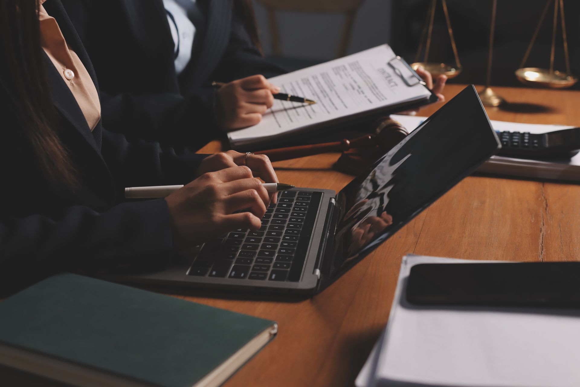 Two people in business suits working with a laptop, documents, and scales of justice on a desk.