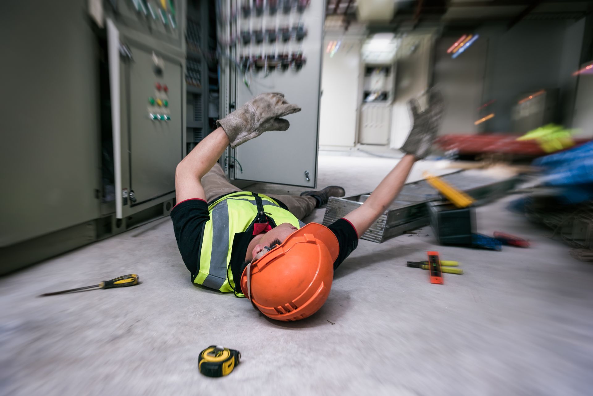 Worker wearing a hard hat and vest has fallen on the floor near electrical equipment.