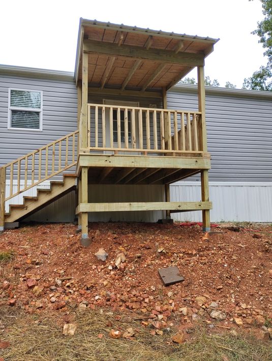 Small wooden porch with stairs beside a gray mobile home, over wood chips.