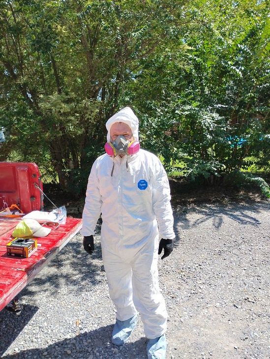 Person in white hazmat suit and respirator stands outdoors near a red table and trees.