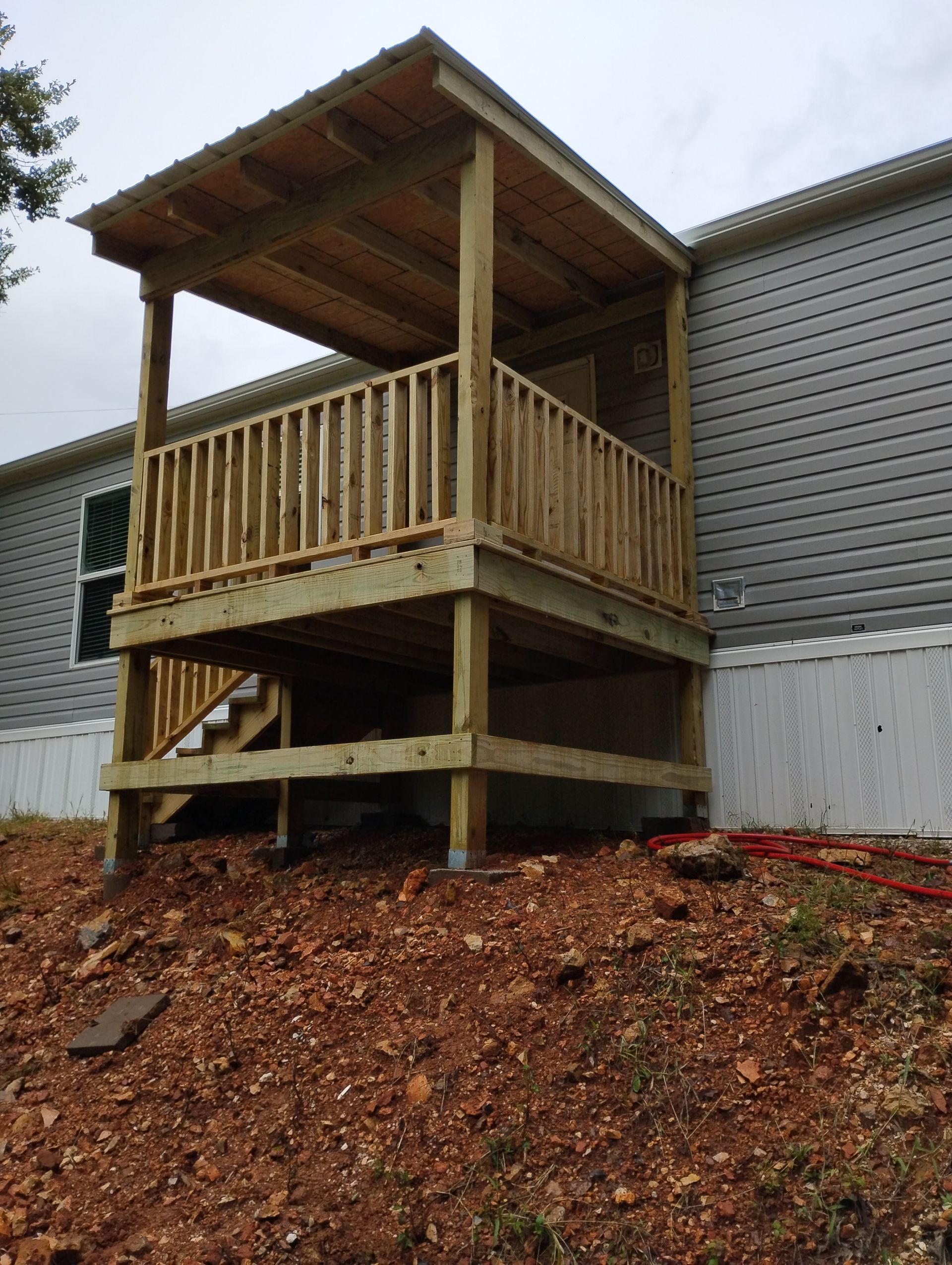 Wooden raised deck with roof and railing beside a gray-sided building, above a mulch-covered slope