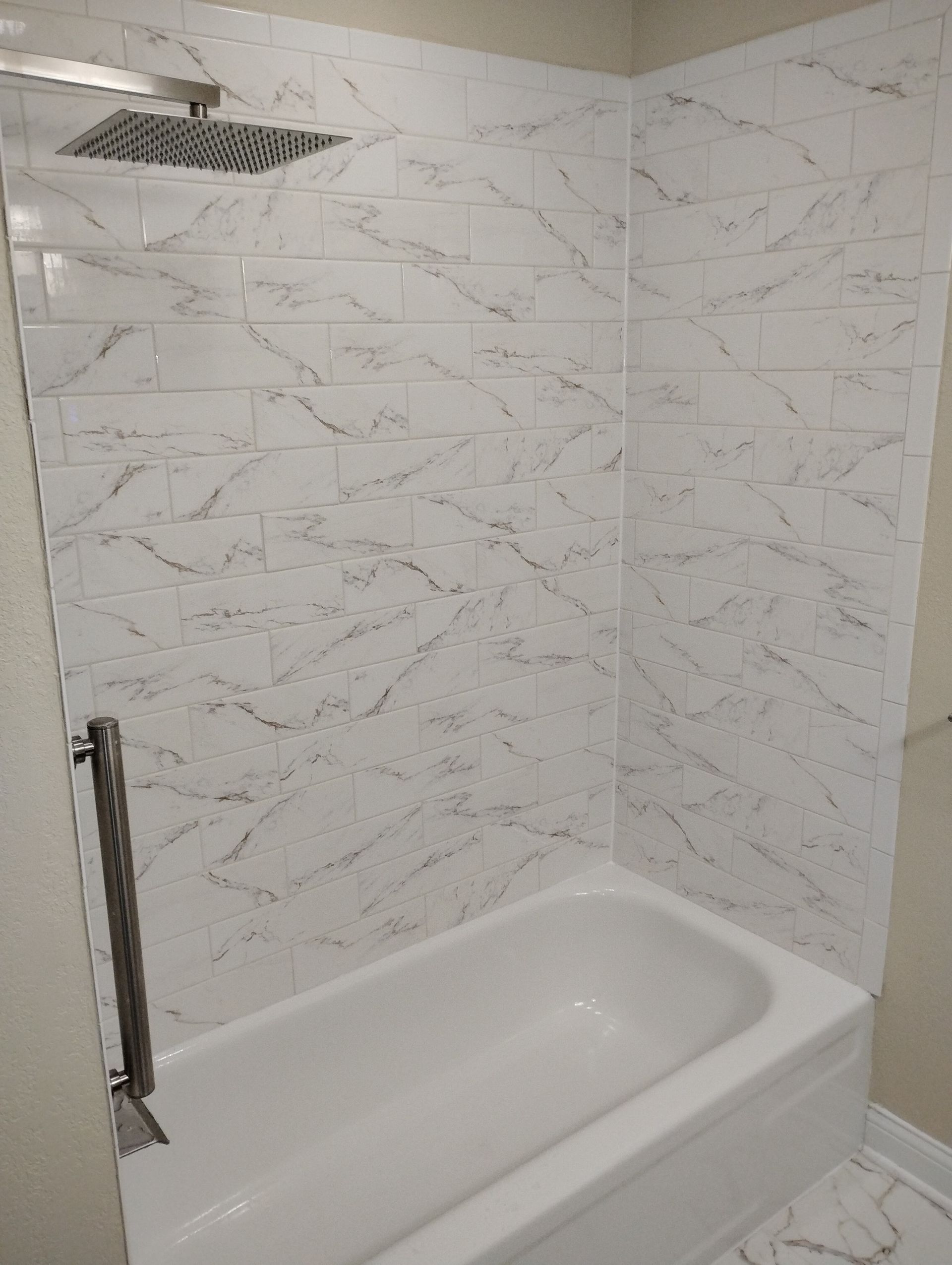 White tiled shower with bathtub, marble-look walls, and a chrome grab bar.
