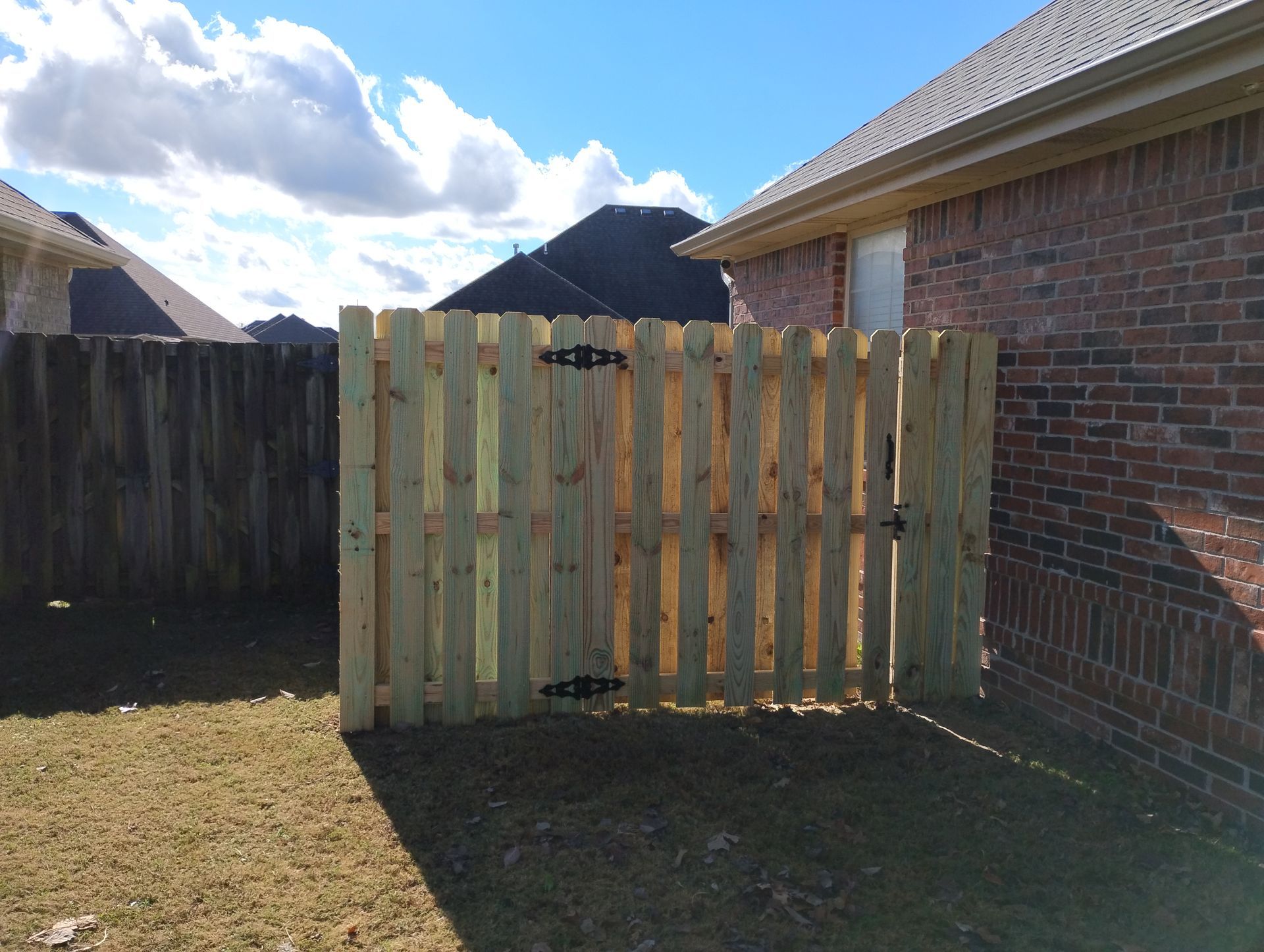 Wooden gate panel leaning beside a brick house in a sunny backyard with a fenced yard.