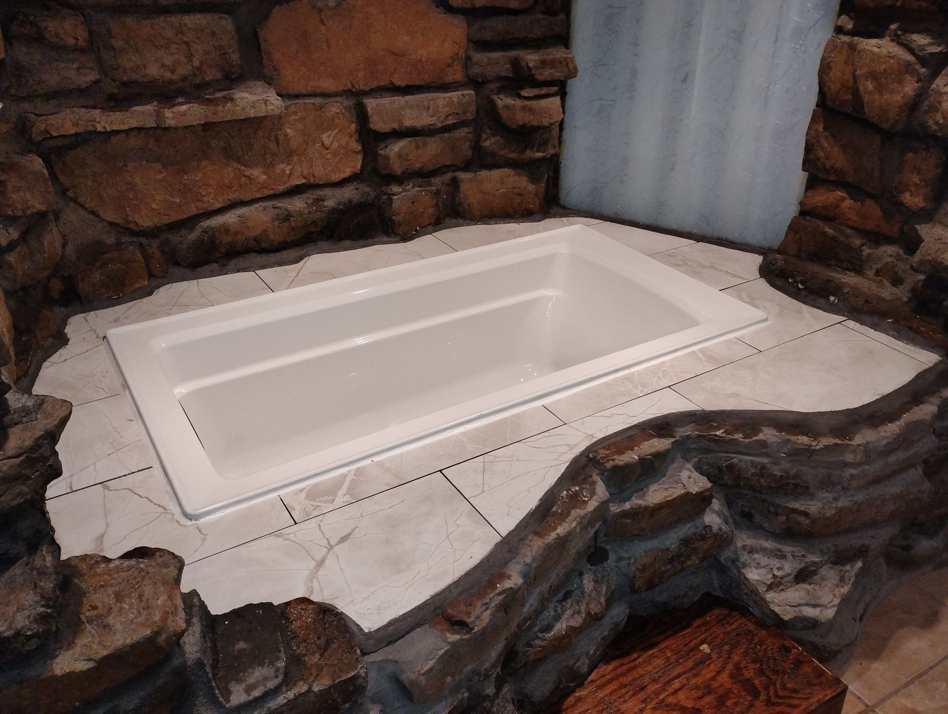 White built-in soaking tub surrounded by stone walls and tiled floor in a rustic bathroom