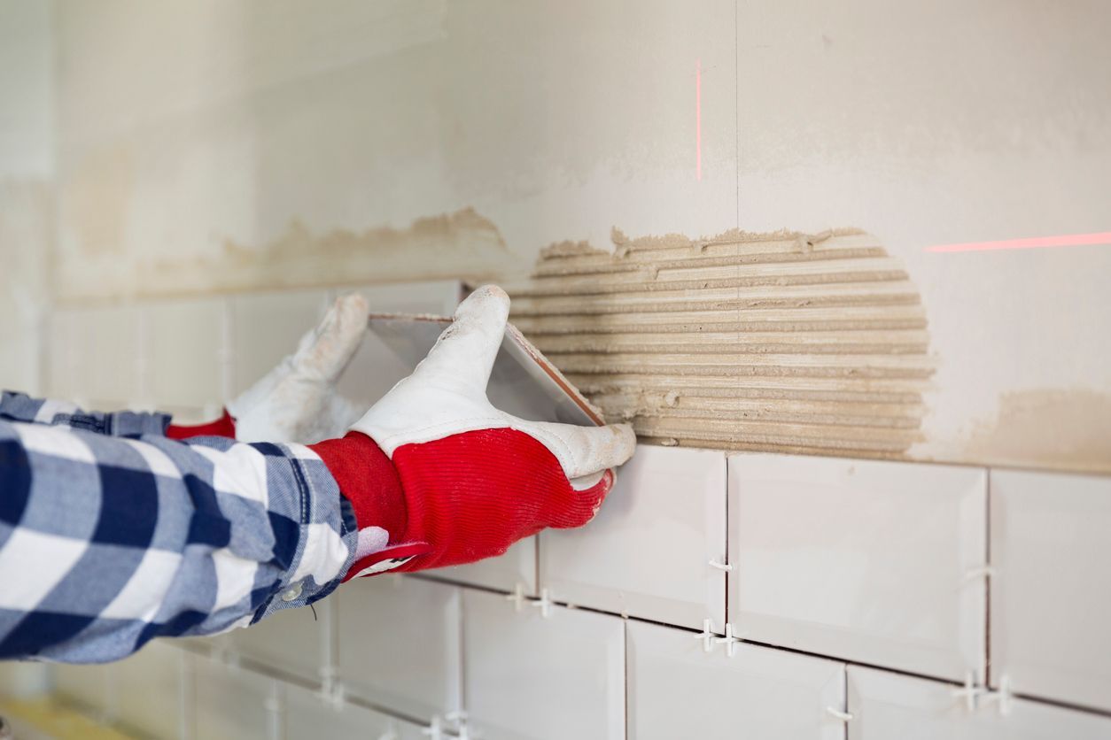 Gloved hand spreading adhesive on a bathroom wall above white tiles
