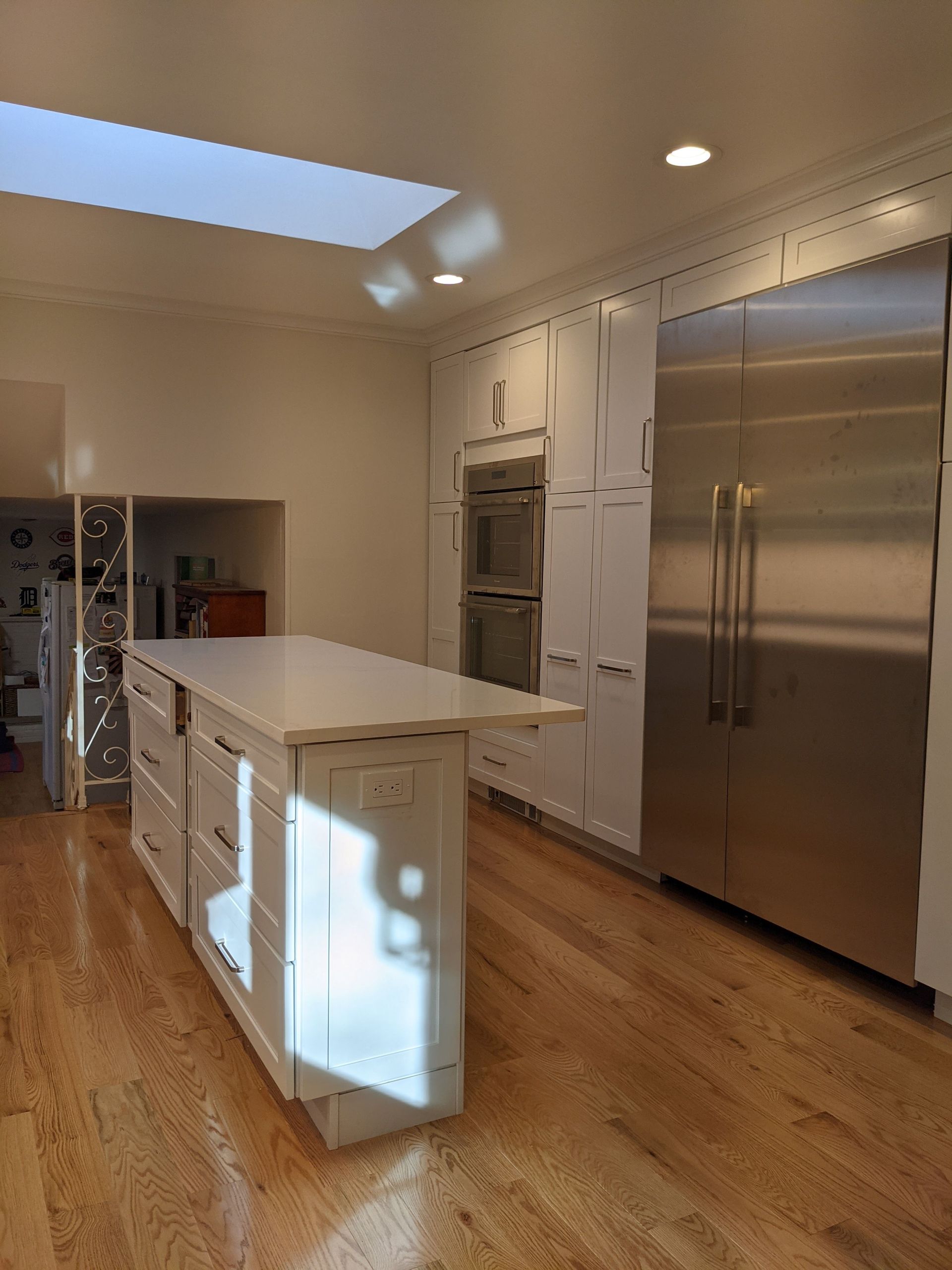 Modern kitchen with white island, stainless steel refrigerator, and wood floors under skylight.