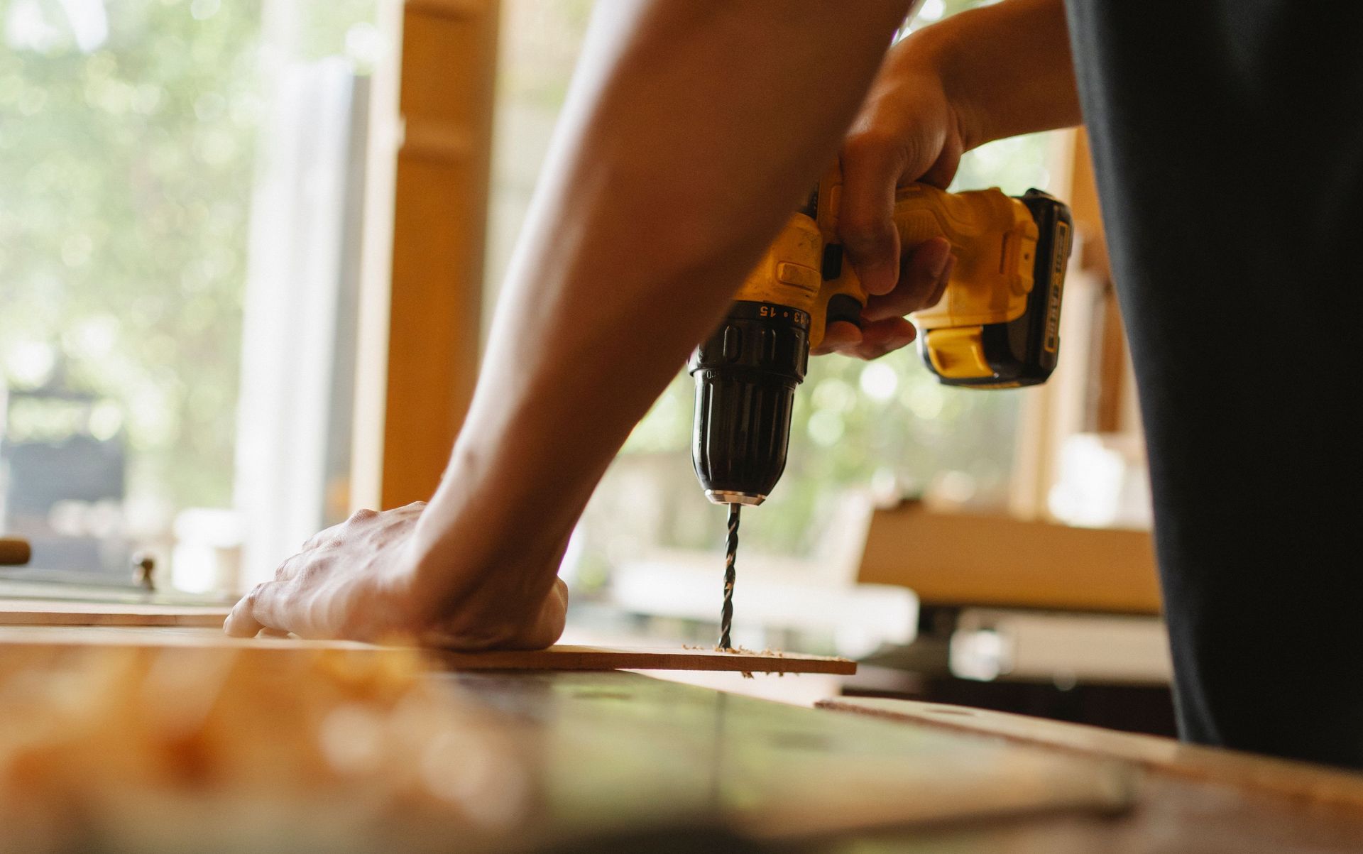 Person drilling a hole in a wooden plank on a workbench near a window
