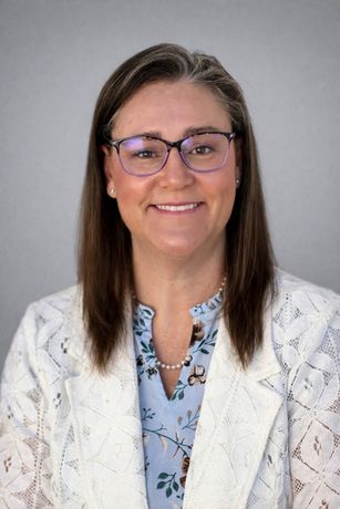 Smiling woman in glasses wearing a white blazer and floral blouse against a gray background