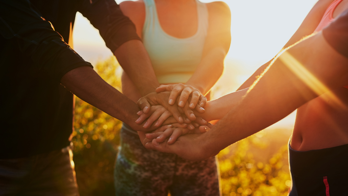Hands stacked together outdoors at sunset, with warm sunlight and athletes in tank tops.