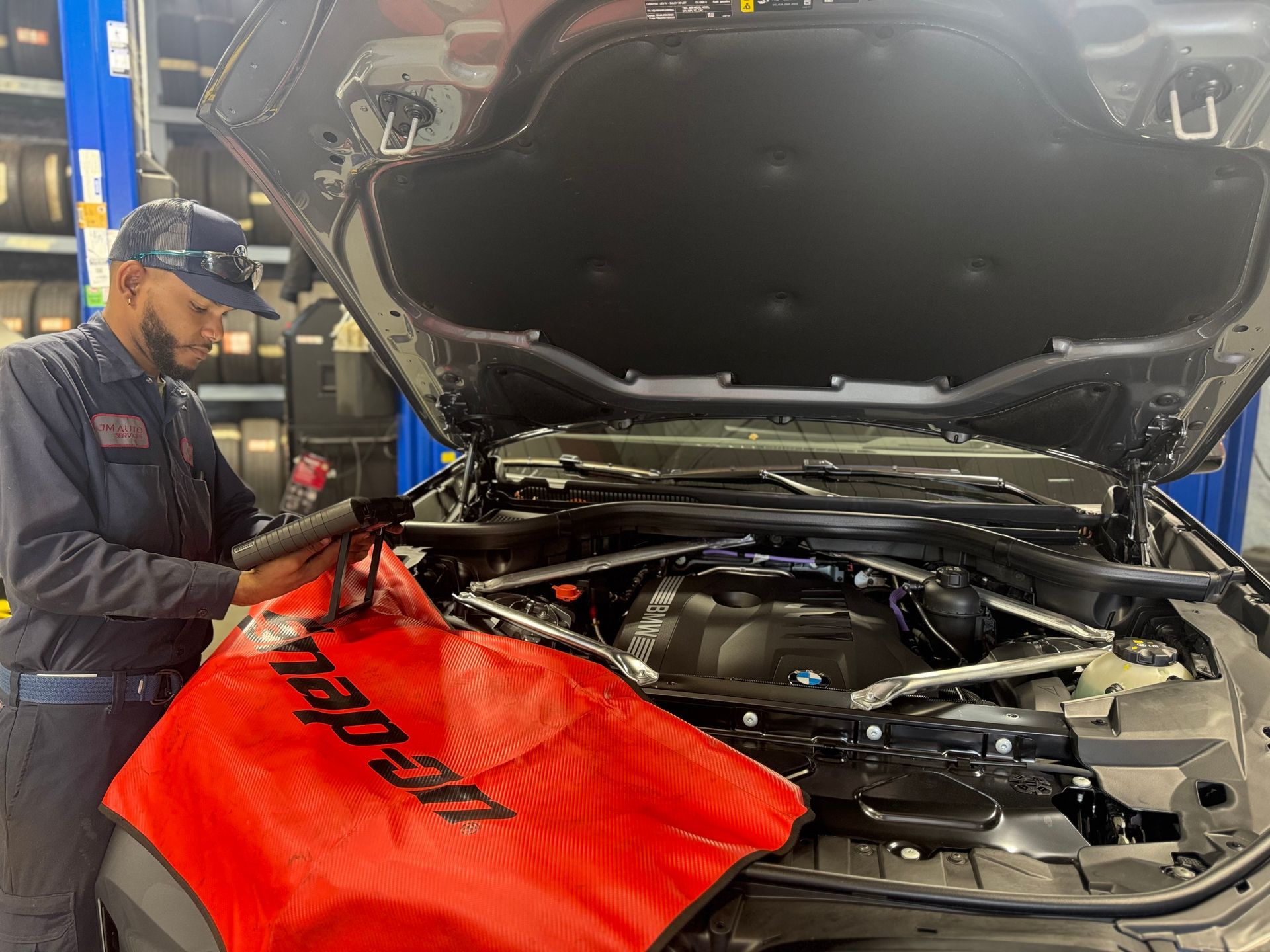 Mechanic working on a black BMW engine with the hood open in a shop, using a red snap-on mat.