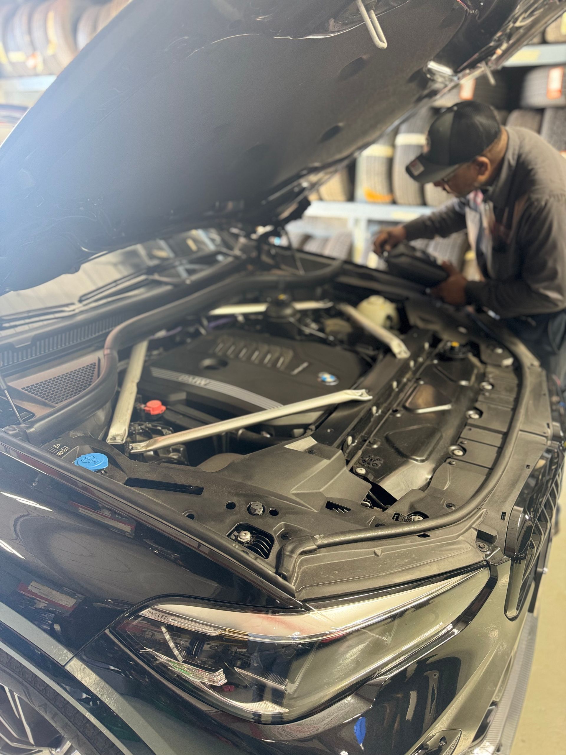A mechanic works on a black car with the hood open. The setting is a garage.